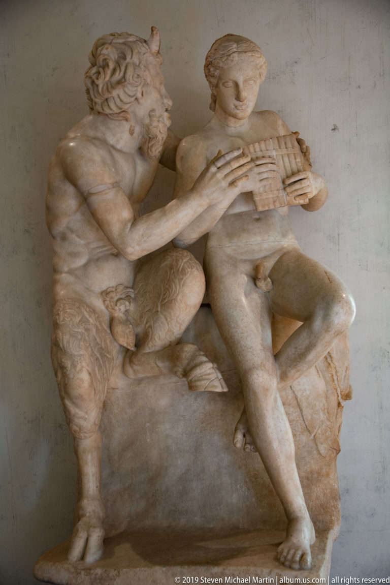 Marble group of goat-legged Pan leaning toward a seated youth who holds a set of pipes.