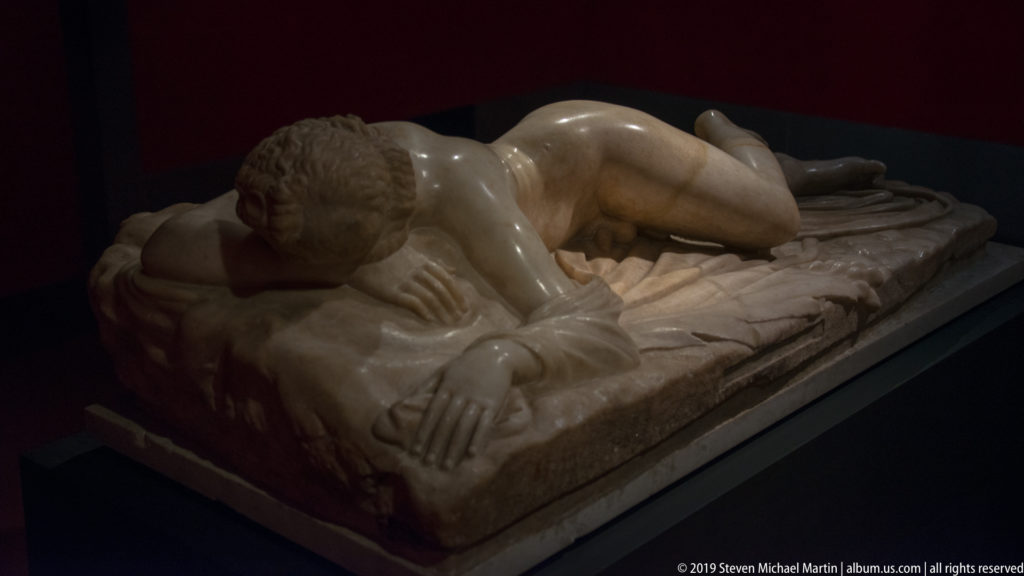 White marble youth lying face-down on carved drapery, head resting on folded arms in a dark display.
