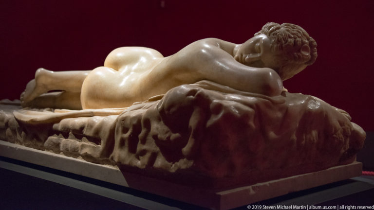 Reclining nude marble figure lying face down on a deeply carved mattress against a dark red wall.
