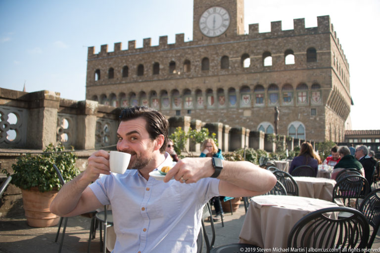 Man sipping from a white cup at a terrace table with café seating and Palazzo Vecchio behind.