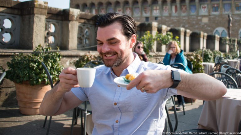 Man holding a white coffee cup and saucer with pastry at an outdoor café, Palazzo Vecchio behind.