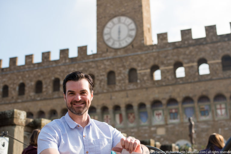 Smiling seated man with Palazzo Vecchio clock, crenellations, arches, and painted shields behind him.