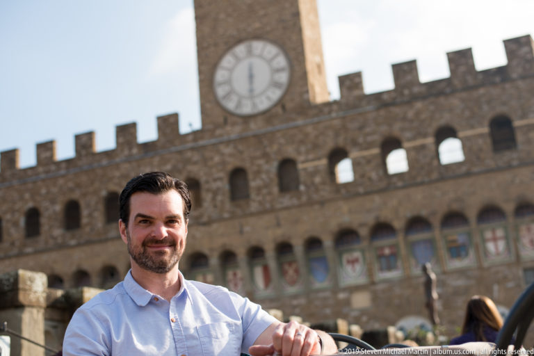 Seated man on an outdoor terrace with the clock and crenellated façade of Palazzo Vecchio behind.