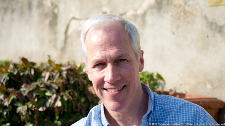 Close portrait of a smiling gray-haired man outdoors beside leafy plants and a pale wall.