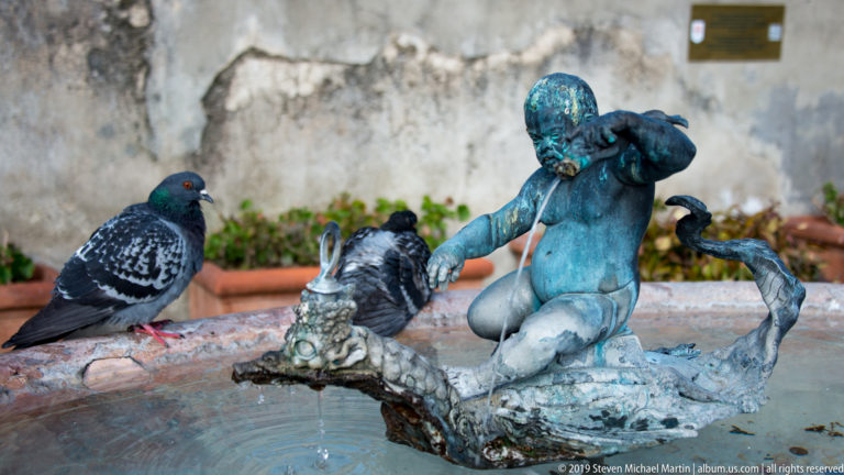 Blue-green patinated fountain figure in a shallow basin with pigeons perched around the sculpture.