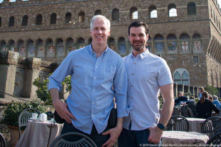 Two men standing beside café tables with the crenellated Palazzo Vecchio façade behind them.