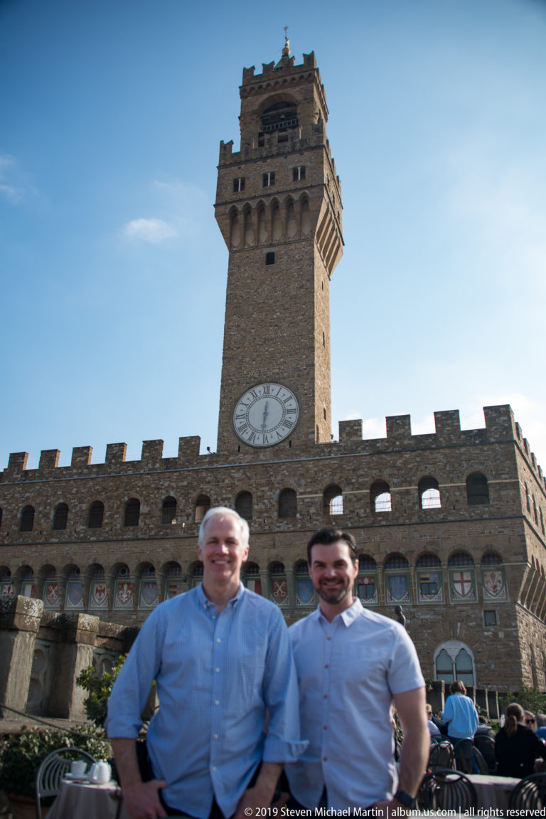 Two men posing together outdoors with Palazzo Vecchio and the Arnolfo Tower behind them.