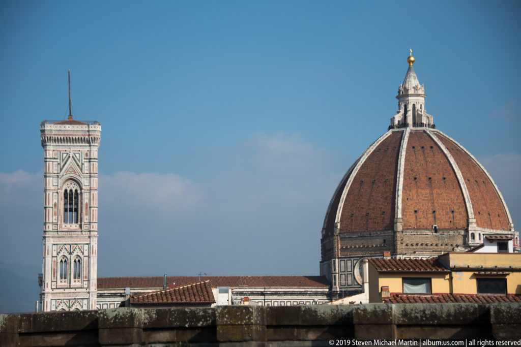 Florence Cathedral dome and Giotto Campanile rising above tiled rooftops under a blue sky.