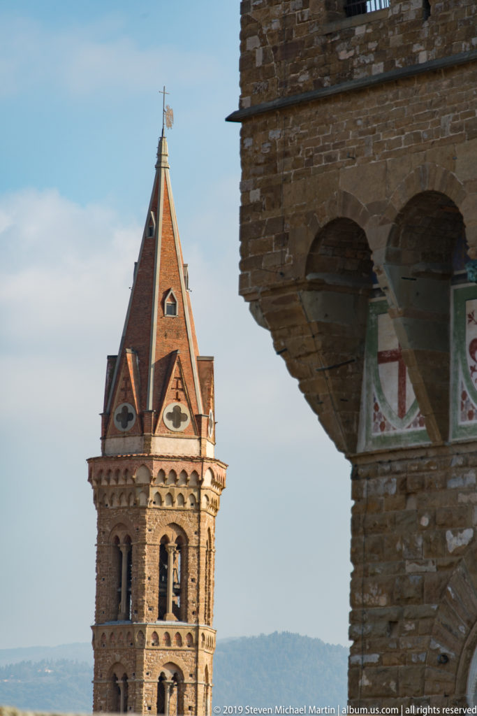 Slender Gothic bell tower with red pointed roof beside the edge of a rusticated stone building.