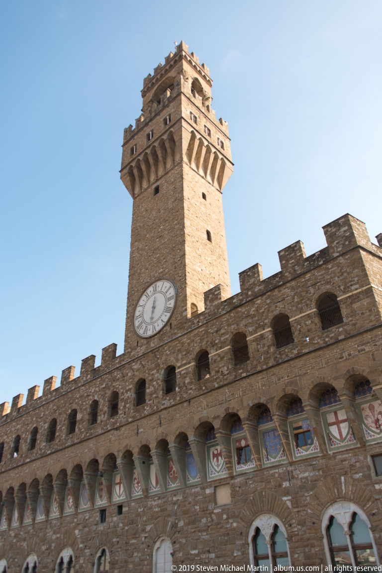 Low-angle view of Palazzo Vecchio with Arnolfo Tower, clock, crenellations, arched openings, and painted shields.