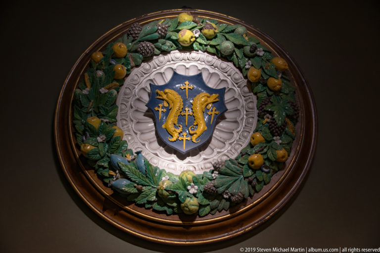 Circular glazed ceramic heraldic relief with blue shield, golden creatures, and a wreath of green leaves and fruit.