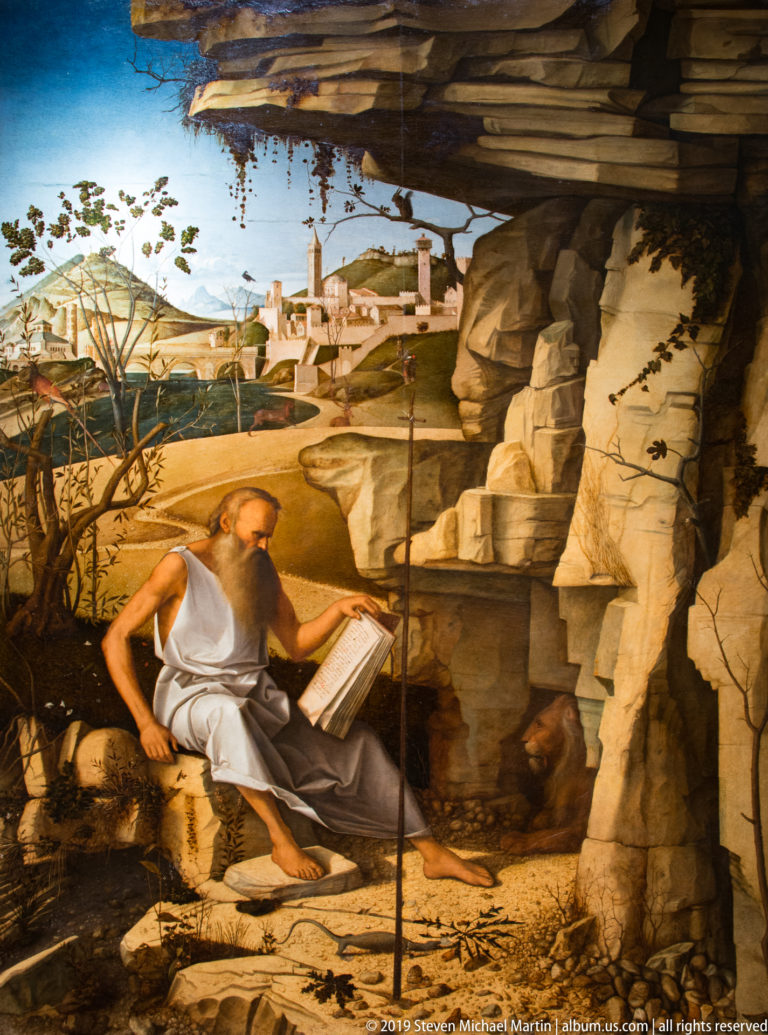 Bearded draped hermit reading an open book among rocks, with a distant city in the landscape.