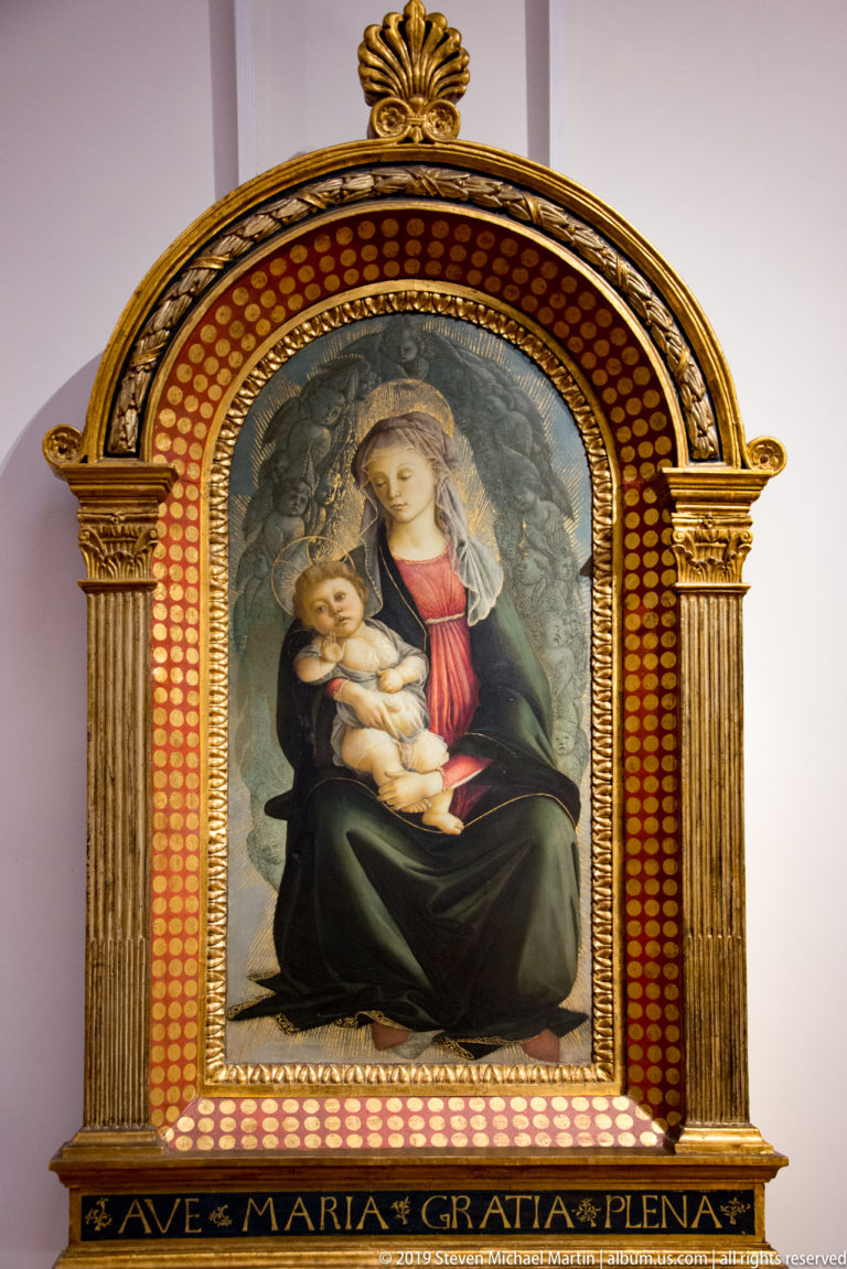 Virgin in green and red cradles the infant Christ within a gilded frame inscribed AVE MARIA GRATIA PLENA.