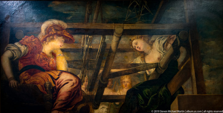 Two women seated beside a large wooden loom or textile frame in a dark interior.