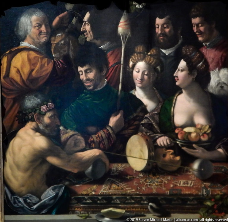 Crowded group of richly dressed men and women with a hand drum, fruit, flowers, and musicians in a dark interior.