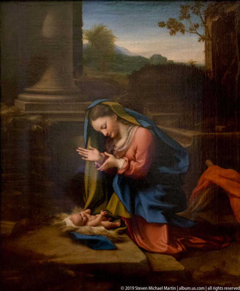 Kneeling Virgin with hands joined beside the infant Christ lying on a cloth in a dark landscape.