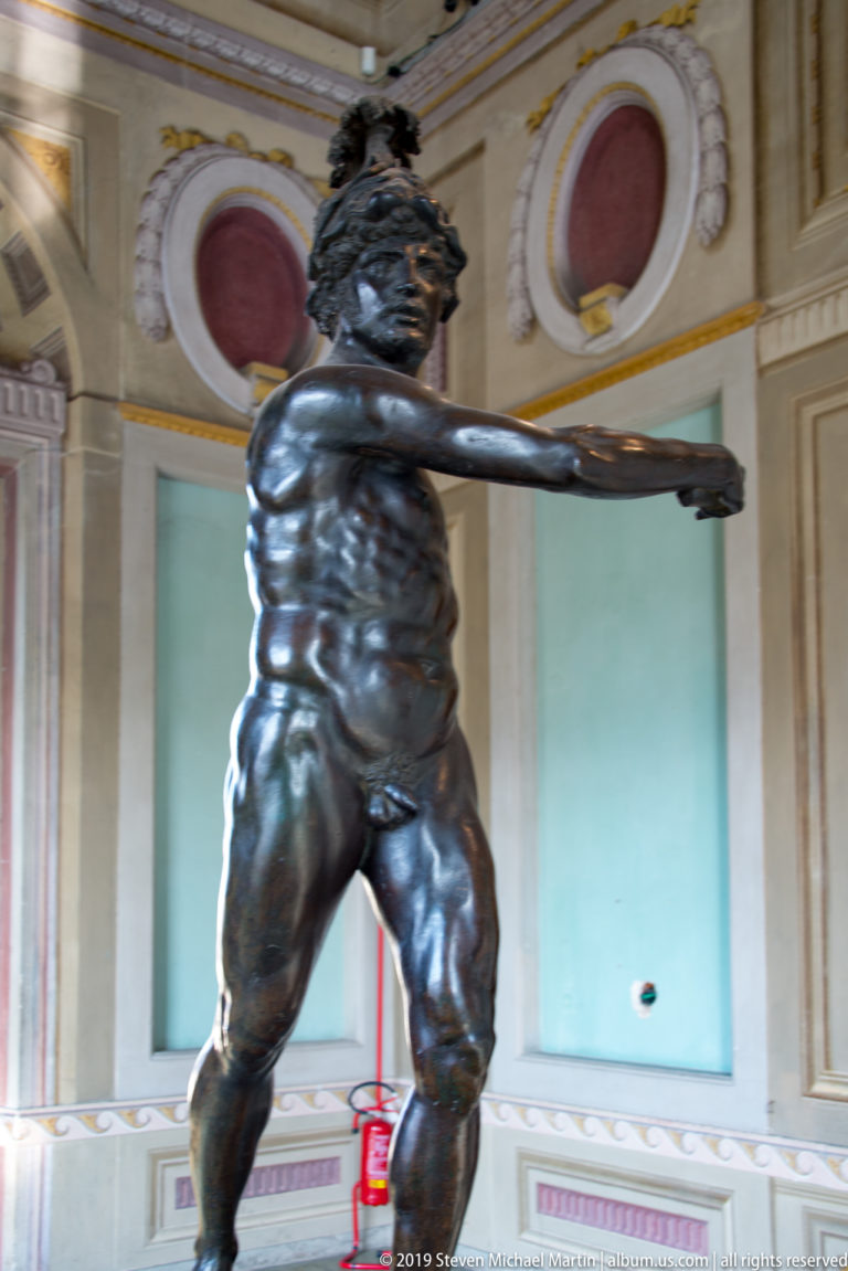Dark bronze nude male statue standing with one arm extended horizontally in a pale museum room.