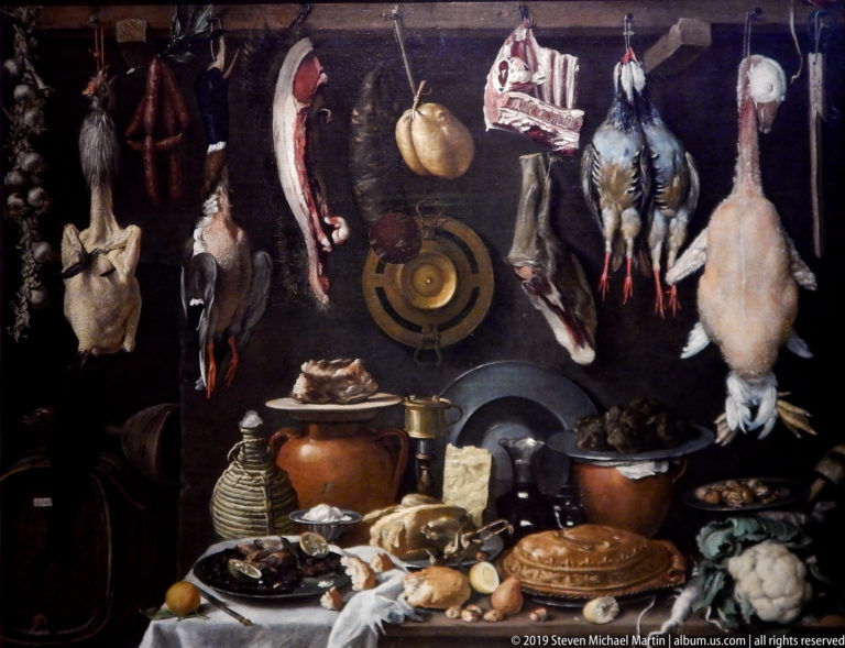 Dark pantry still life packed with hanging birds and meat above pots, bread, cheese, vegetables, and dishes.
