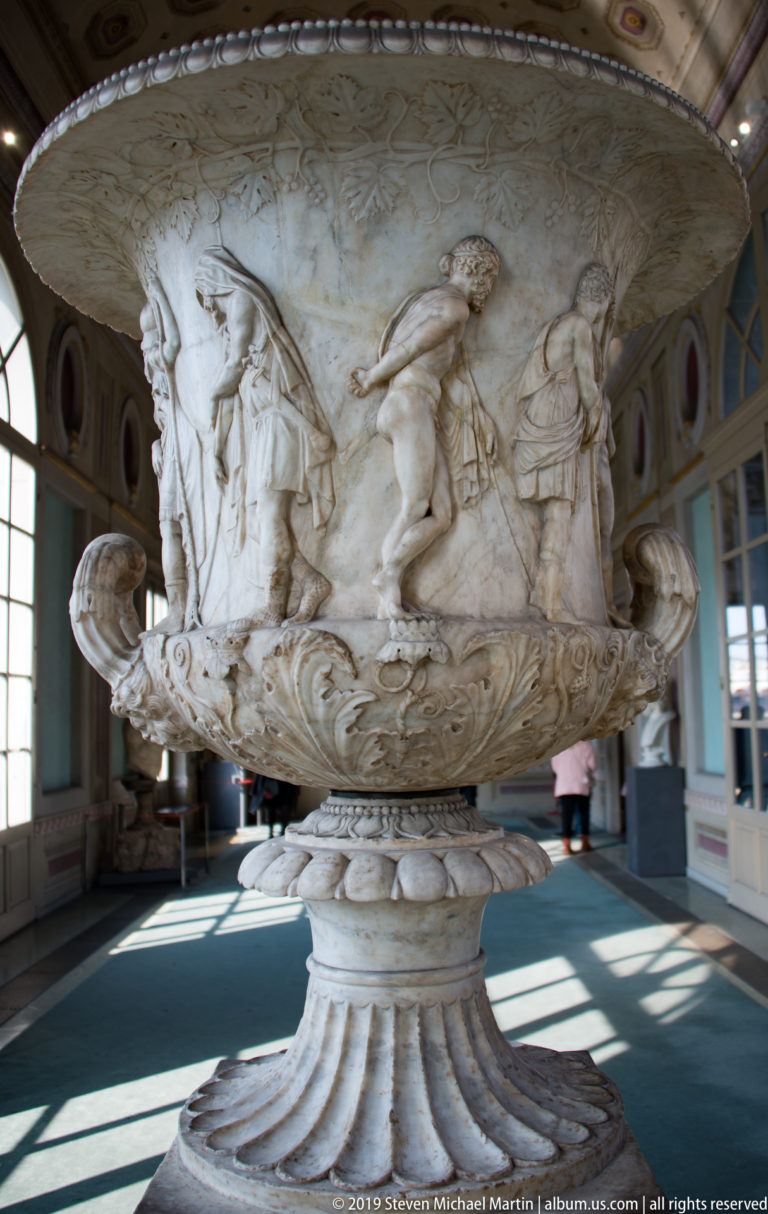 Large white marble vase on a carved pedestal, with relief figures around the bowl and scrolling handles in a gallery.