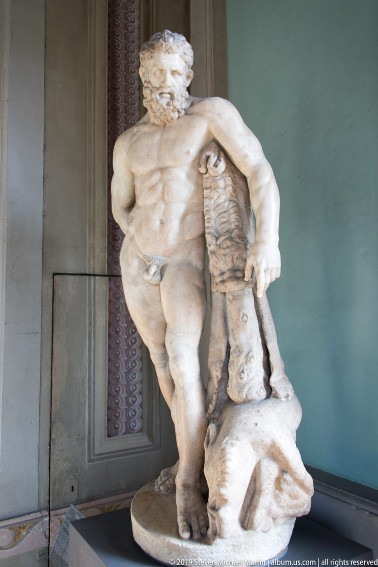 White marble nude male statue leaning beside a support draped with a carved animal skin in a museum gallery.
