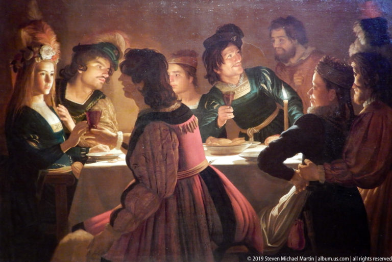 Crowded group of men and women around a brightly lit table, holding glasses and talking in a dark interior.