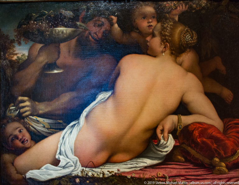 Reclining nude woman seen from behind, surrounded by satyr-like men and small children against dark foliage and red cloth.