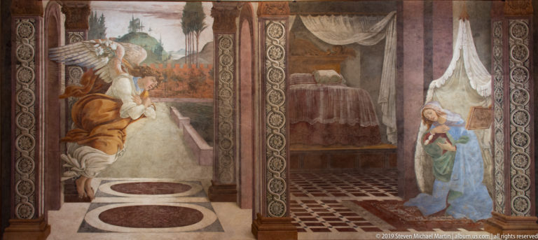 Kneeling angel greets the Virgin in a patterned Renaissance interior with a curtained bed visible in the adjoining room.