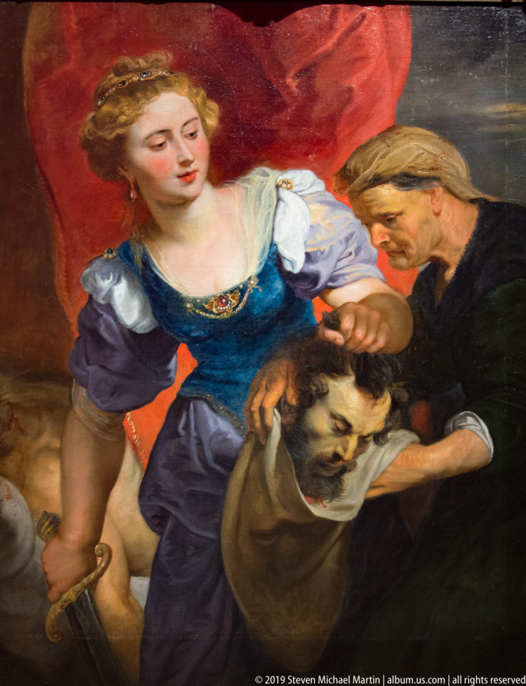 Woman in blue holding a sword beside an older man as they handle the severed head of a bearded man.