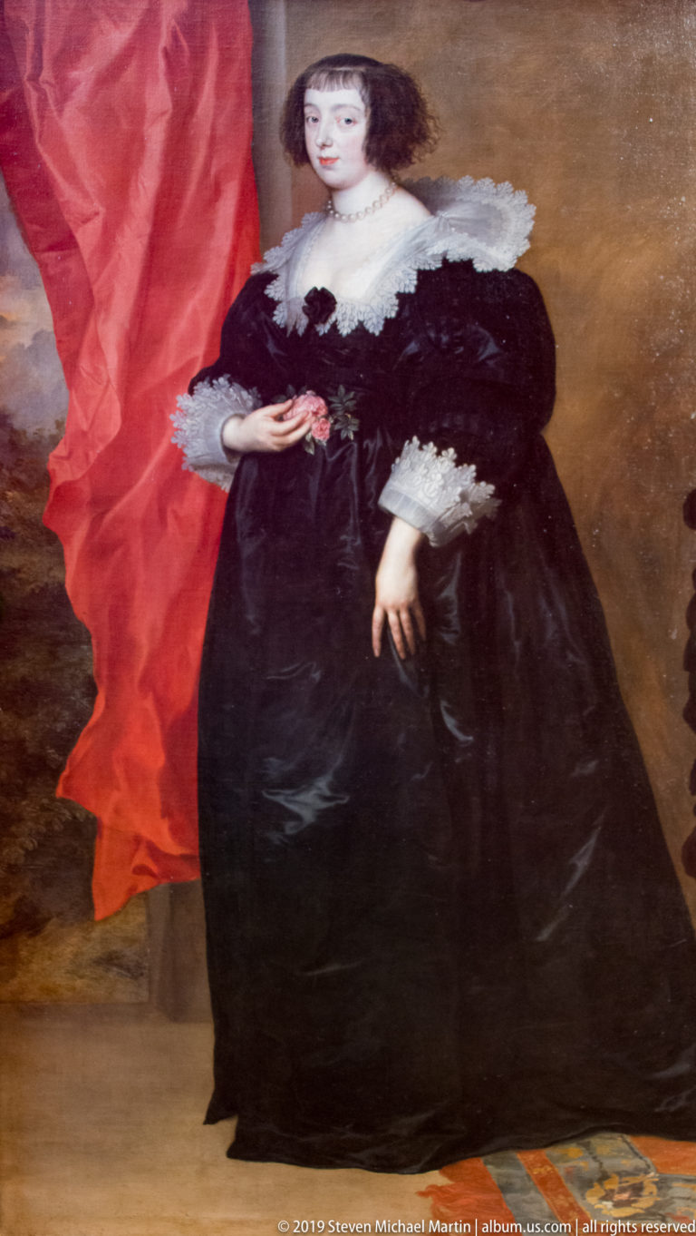 Full-length woman in a black gown with broad white lace collar and cuffs, standing beside red drapery.