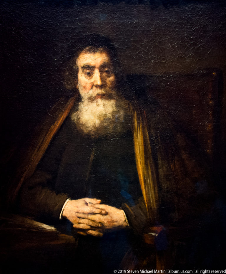 Elderly white-bearded man seated in dark clothing with his hands folded against a brown-black background.
