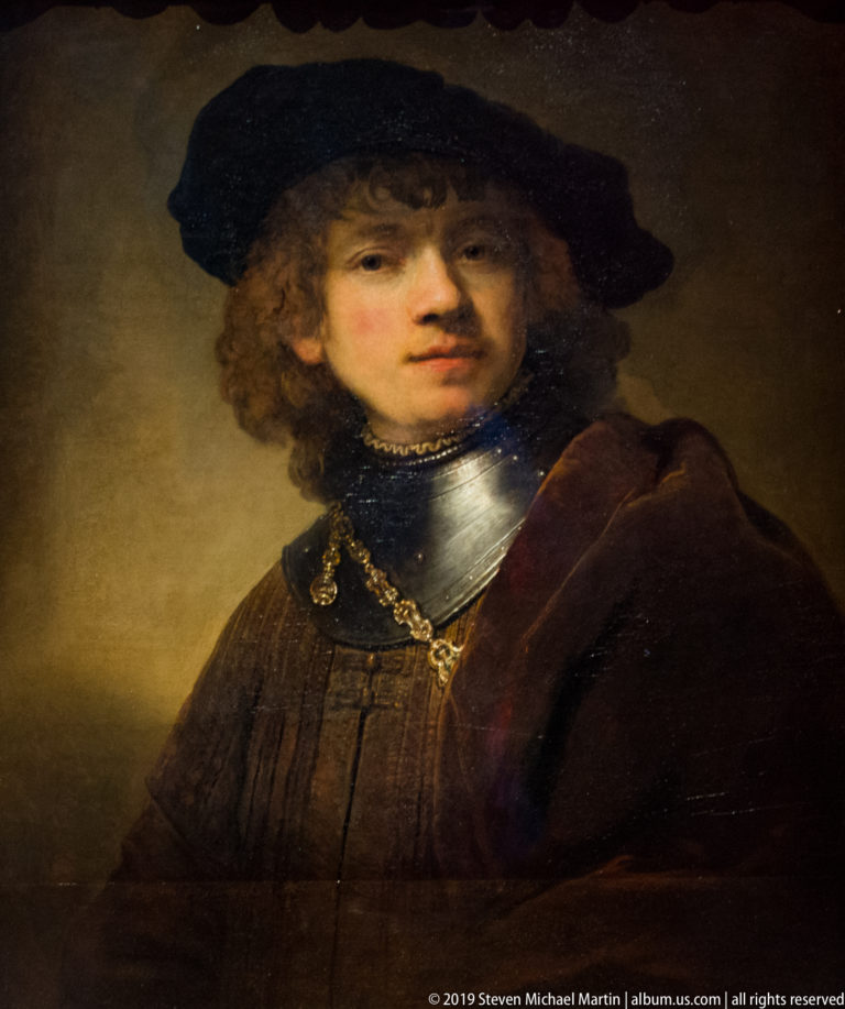 Curly-haired young man wearing a wide dark hat, metal neck armor, brown cloak, and gold chain.