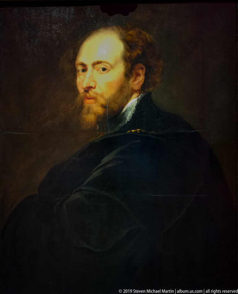 Bearded man in a black cloak turning toward the viewer against a dark brown background.