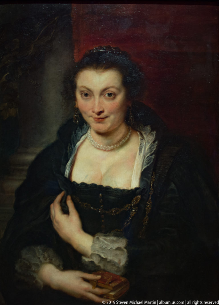 Woman in a black gown with pearl necklace, white lace cuffs, and gold chain seated against a dark red background.