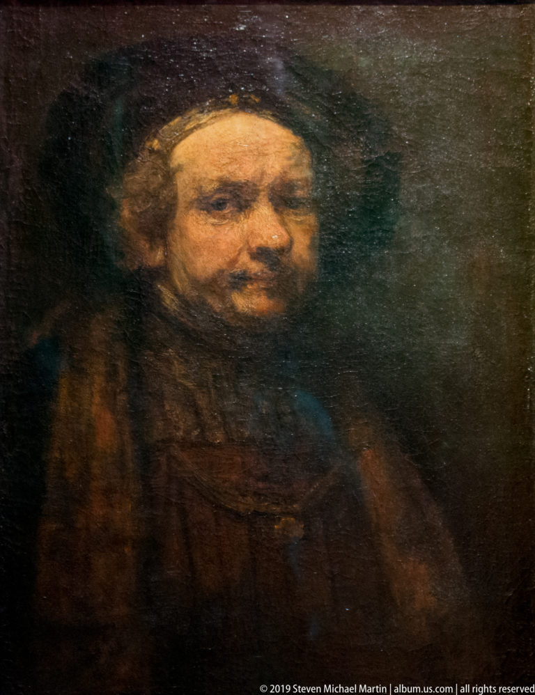 Dark half-length portrait of a bearded man facing forward, his face softly lit against a nearly black background.