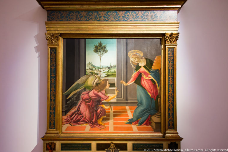 Kneeling angel reaches toward the bending Virgin across a red-tiled interior with a distant landscape visible through a window.