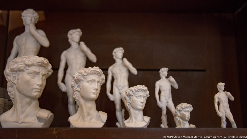 Rows of white marble portrait busts in front of small standing male statues inside a museum display.