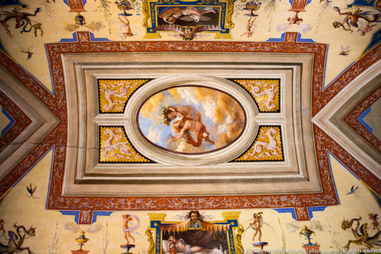 Ornate ceiling centered on an oval painted figure, surrounded by colorful geometric borders and small grotesque figures.
