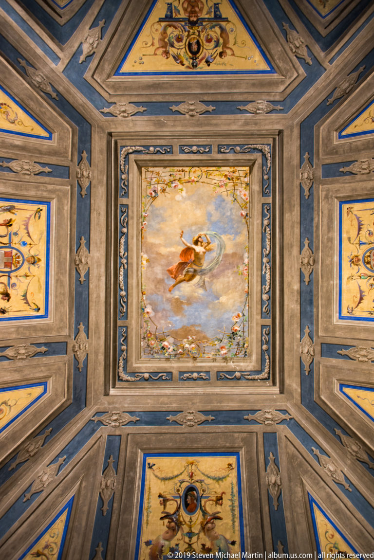 Ornate painted ceiling with a central airborne figure, blue-and-gold panels, grotesque decoration, and molded frames.