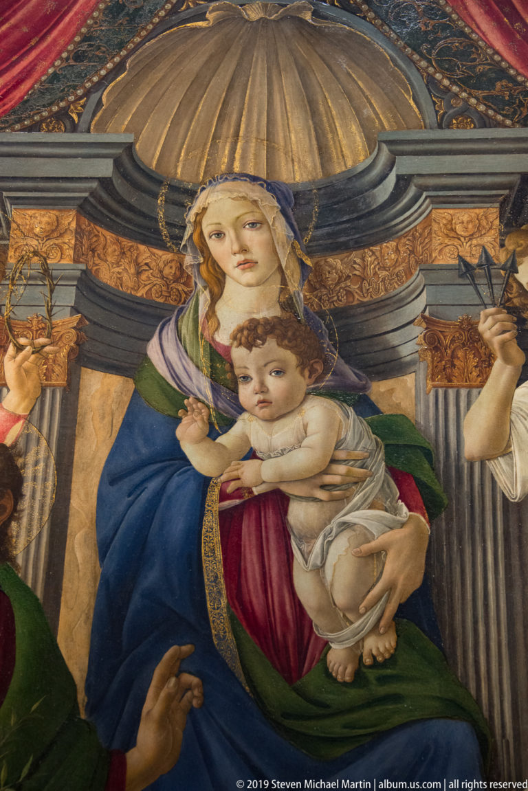 Virgin in blue and red holds the nude Christ Child beneath monumental architecture, with cropped saints at both edges.