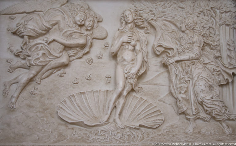 Pale relief of Venus standing above a scallop shell, flanked by wind-borne figures and a draped attendant.