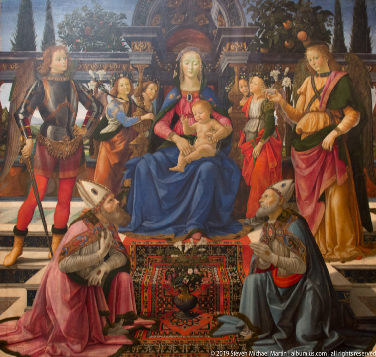 Enthroned Virgin and Child surrounded by angels and saints, with two kneeling figures beside a patterned carpet.