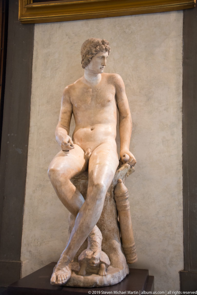 Seated nude marble youth crosses one leg over the other on a rocky support and extends one hand.