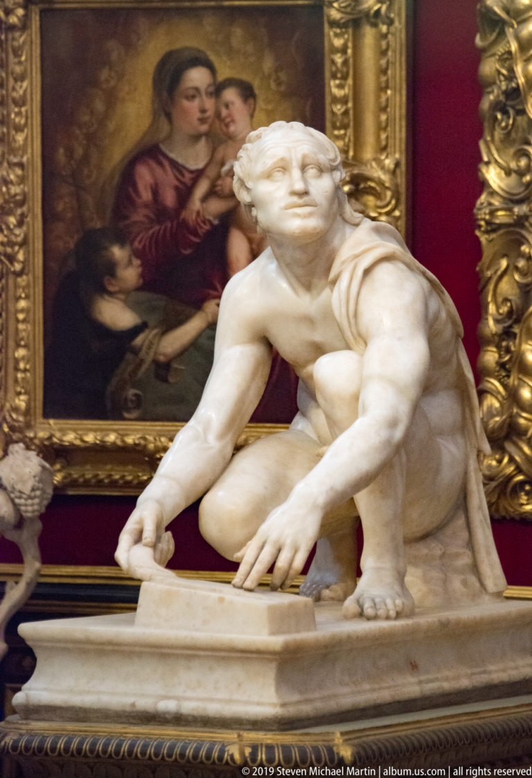 Crouching nude marble male bends over a low rectangular support before a large gilded painting on a red wall.