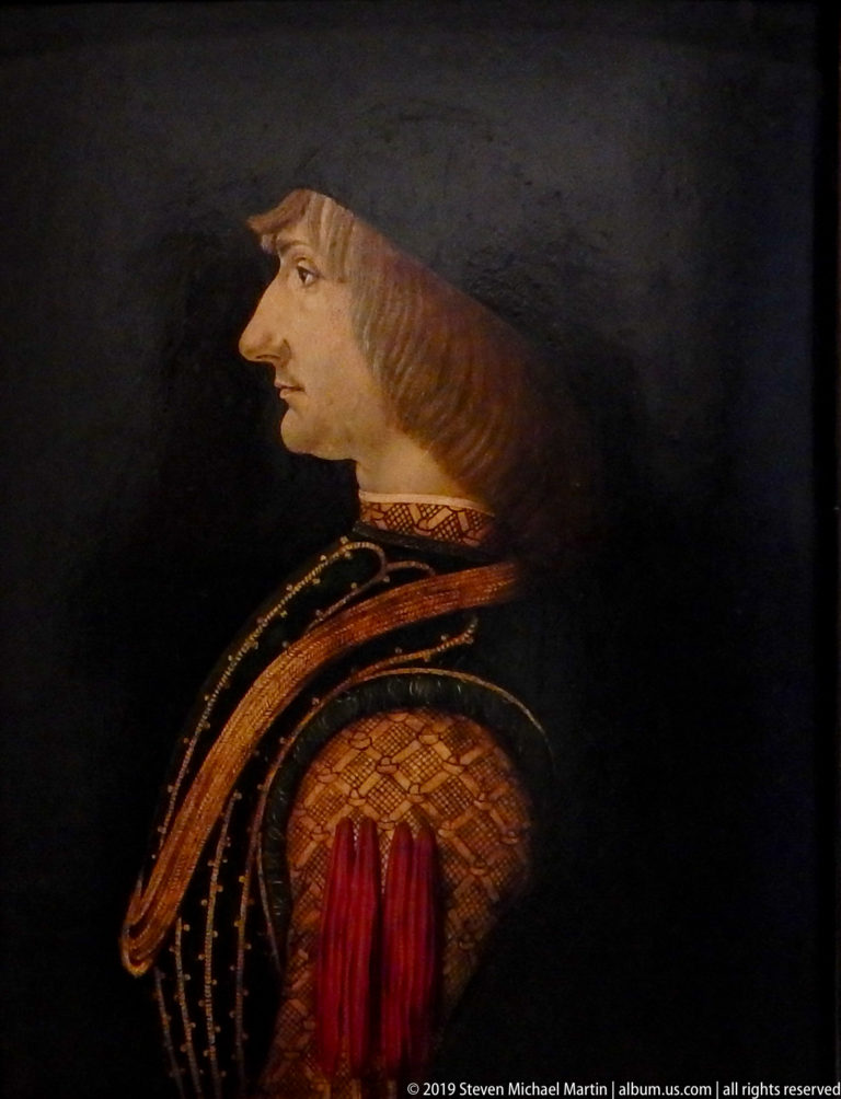 Young man in strict profile with jaw-length brown hair and an ornate red-and-brown patterned garment against black.