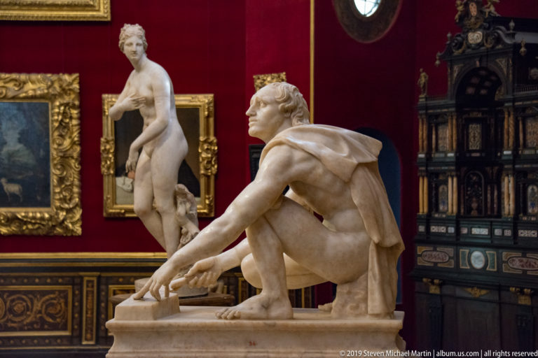 Crouching nude marble male reaches forward with both arms, with a standing statue and tall cabinet behind him.