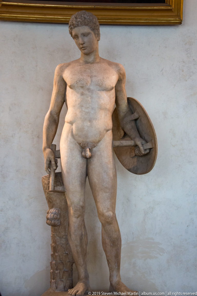 Muscular nude marble male holds a round shield beside a rough tree-trunk support in a museum gallery.