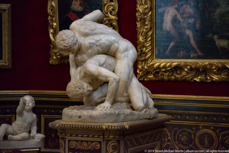 Two nude marble wrestlers grapple on the ground, one crouched over and pinning the other on an ornate pedestal.