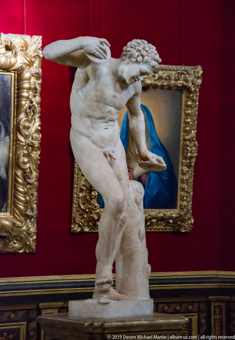 Dynamic nude marble male twists with one arm extended in a red-walled gallery lined with gilded paintings.