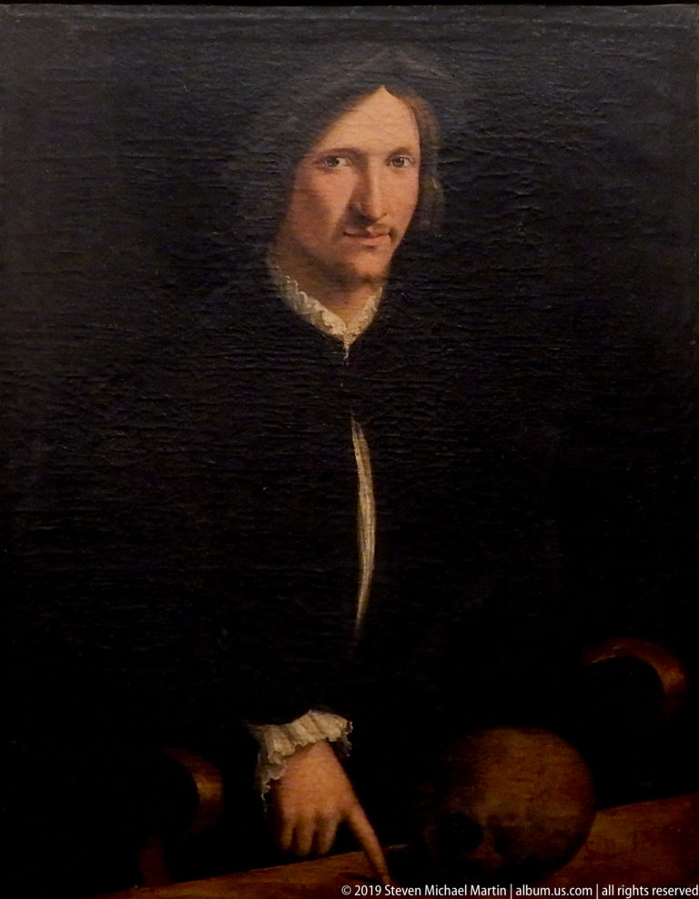 Dark-haired man in black sits against a nearly black background, with one hand visible and a small white collar.