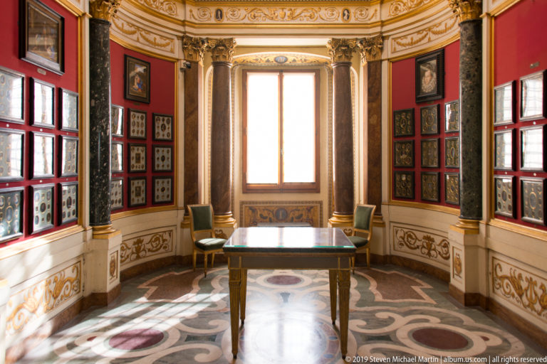 Ornate Uffizi room with red walls, dark columns, small framed works, central window, table, and patterned marble floor.